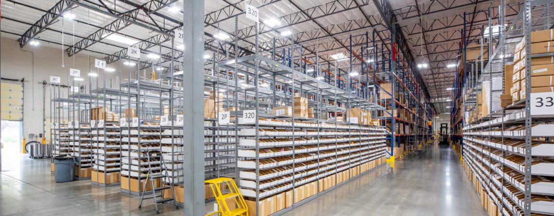 Large, organized warehouse interior with tall metal shelving units filled with boxed inventory, wide polished concrete aisles, overhead industrial lighting, and clearly marked storage locations.